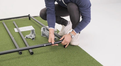 SimBox Golf Simulator Enclosure with a black metal frame being assembled over green turf by a person